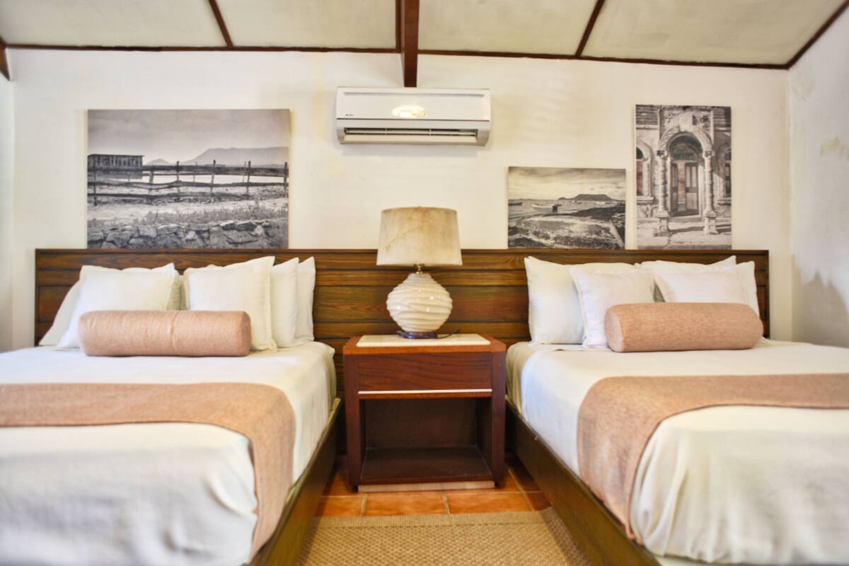 A hotel room with two beds, a wooden headboard, a nightstand with a lamp, and wall art depicting landscapes and architecture.