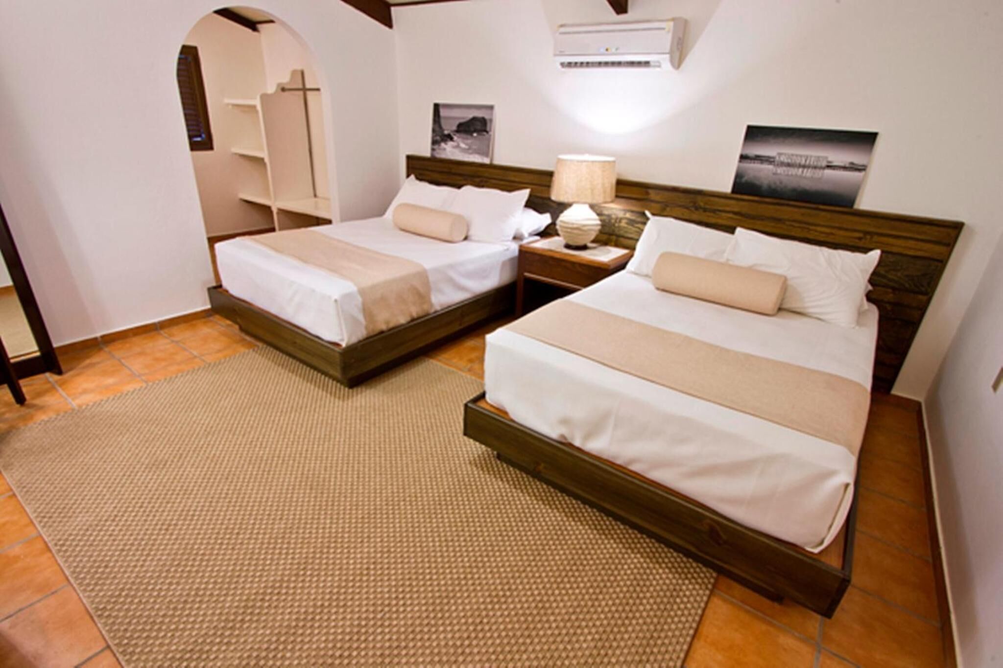 A hotel room with two beds, a small table with a lamp, a wall-mounted air conditioner, and a picture on the wall.