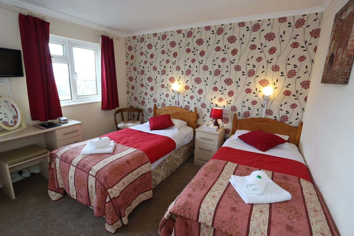 A room with two beds, each with a red and beige bedspread, a wooden headboard, and a nightstand with a lamp.