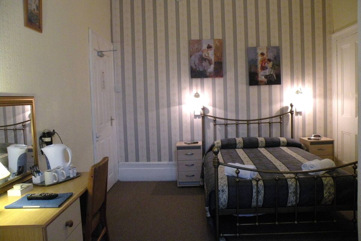 A bedroom with a bed, a desk with a chair, a lamp, and a painting on the wall.