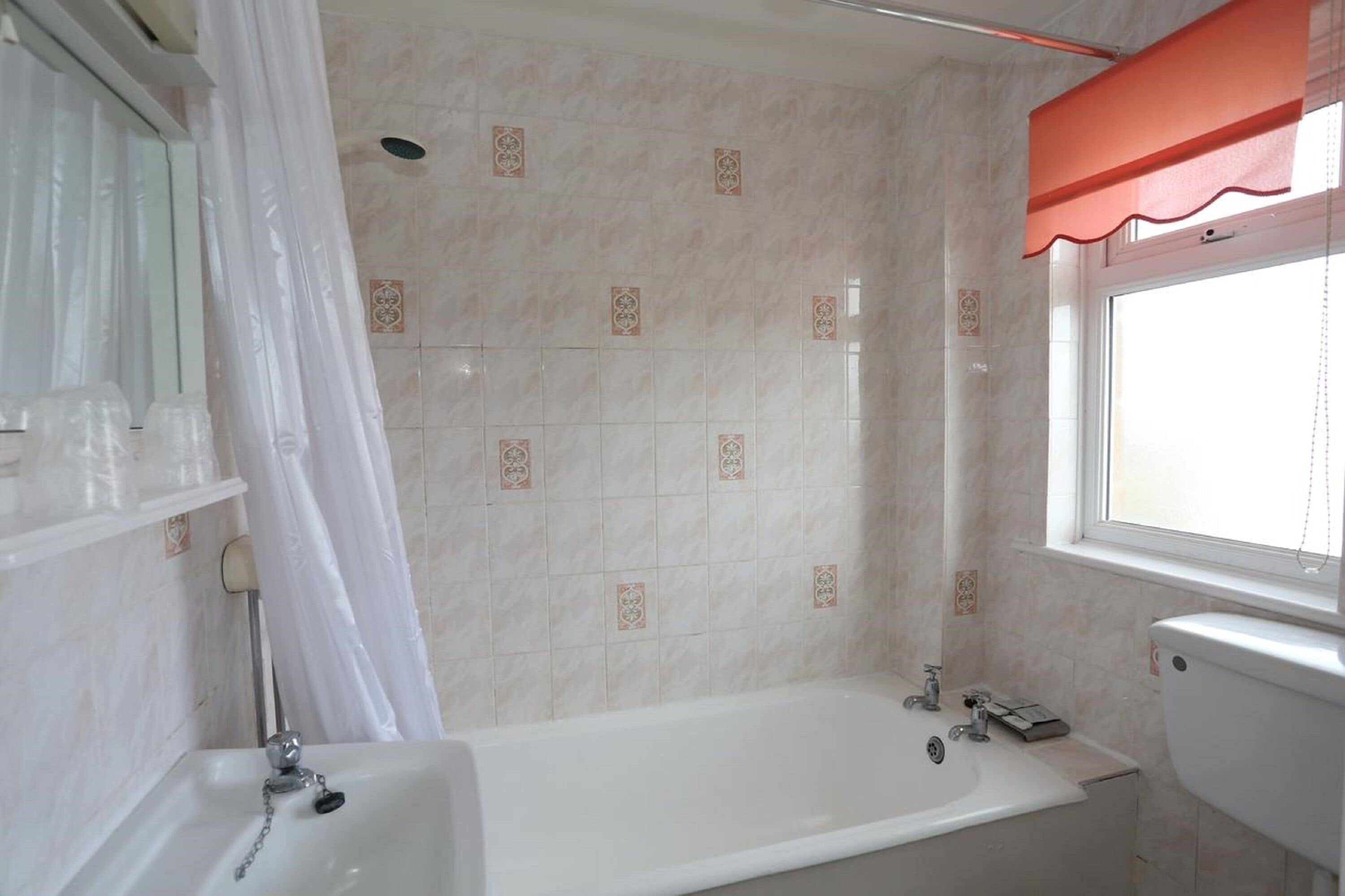Un baño con bañera, inodoro, ventana con persiana roja y cortina de ducha.
