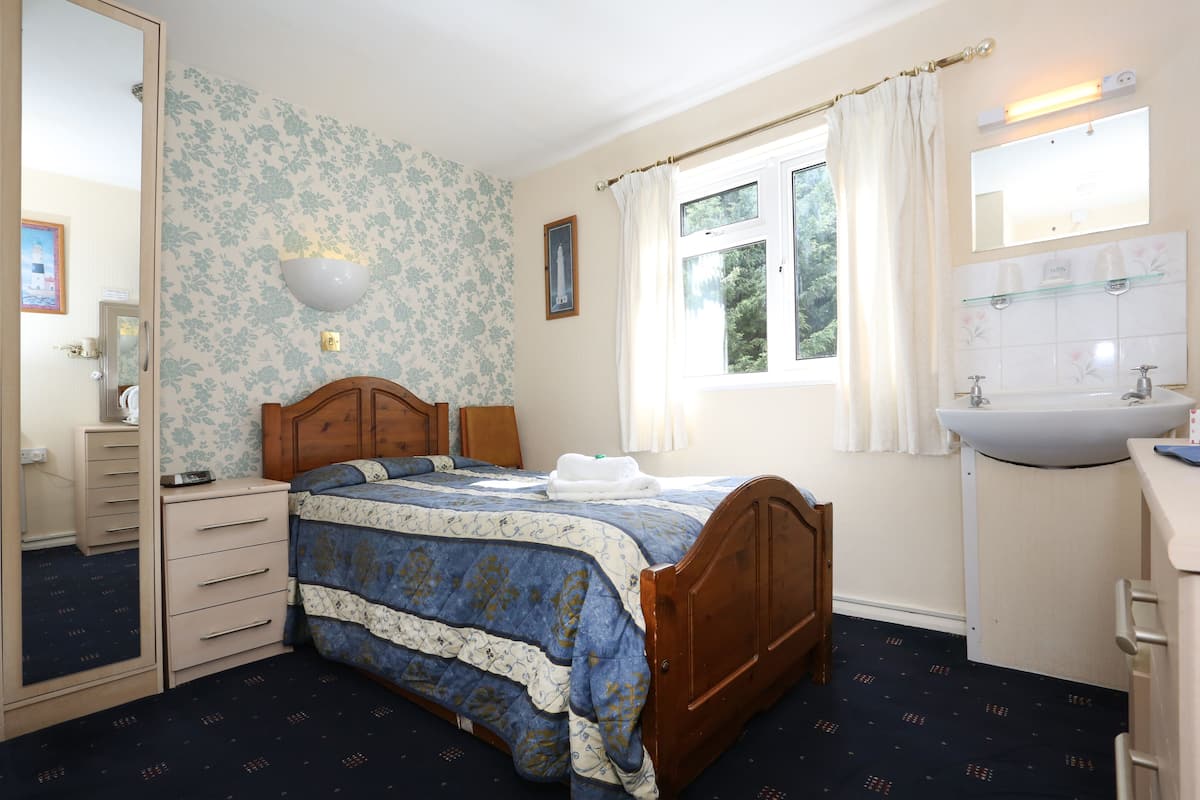 A bedroom with a wooden bed, a dresser, a mirror, a window with curtains, and a bathroom with a sink and mirror.