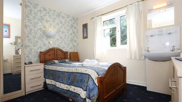 Un dormitorio con cama de madera, cómoda, espejo, ventana con cortinas y baño con lavamanos y espejo.