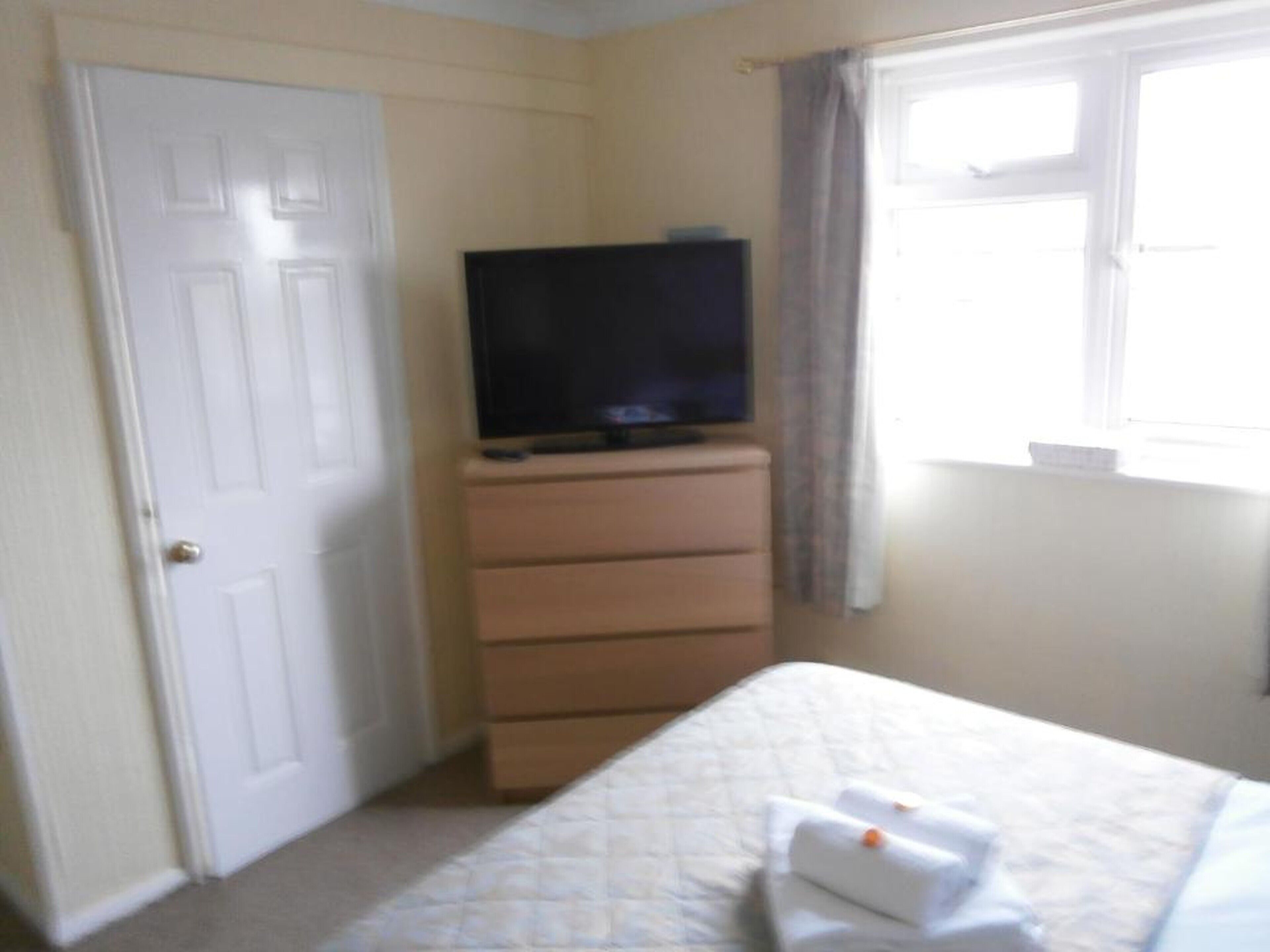 Un dormitorio con cama, cómoda, televisor encima de la cómoda y ventana con cortinas.