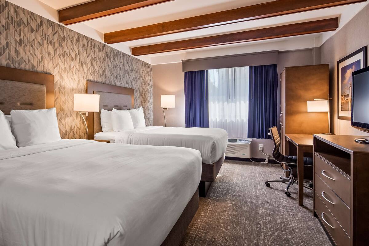 Habitación de hotel con dos camas, un escritorio, una silla, un televisor y un ventanal con cortinas.