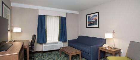 A hotel room with a desk, a blue sofa, a small table, a green chair, a window with blue curtains, and a framed picture on the wall.