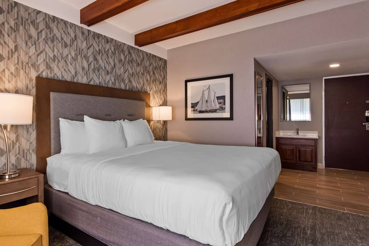 Habitación de hotel con una cama grande, dos lámparas de noche, un cabecero de madera, un cuadro enmarcado en la pared y un baño visible a través de una puerta abierta.