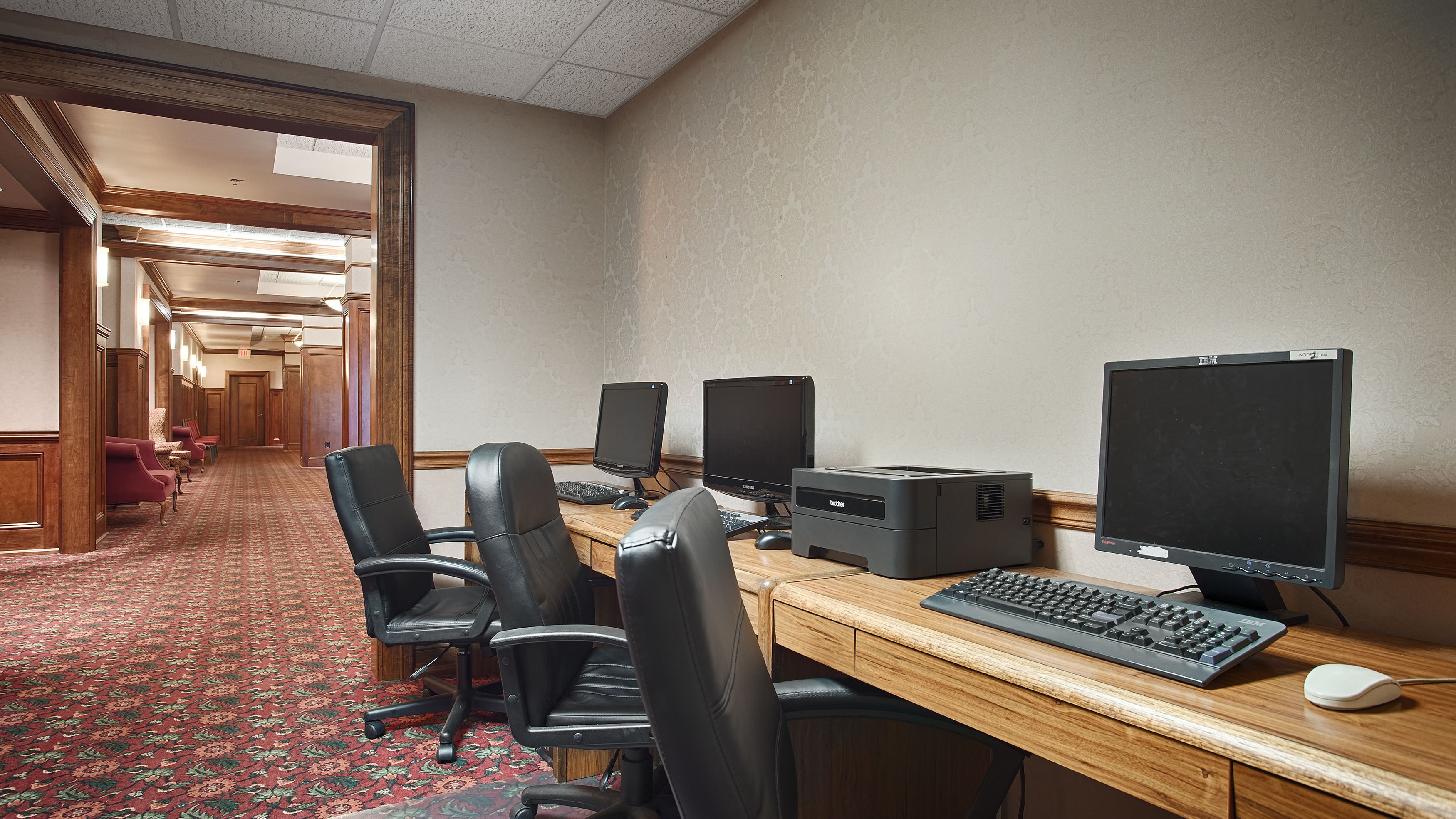 A hotel room with a long desk, multiple monitors, a keyboard, and a printer.