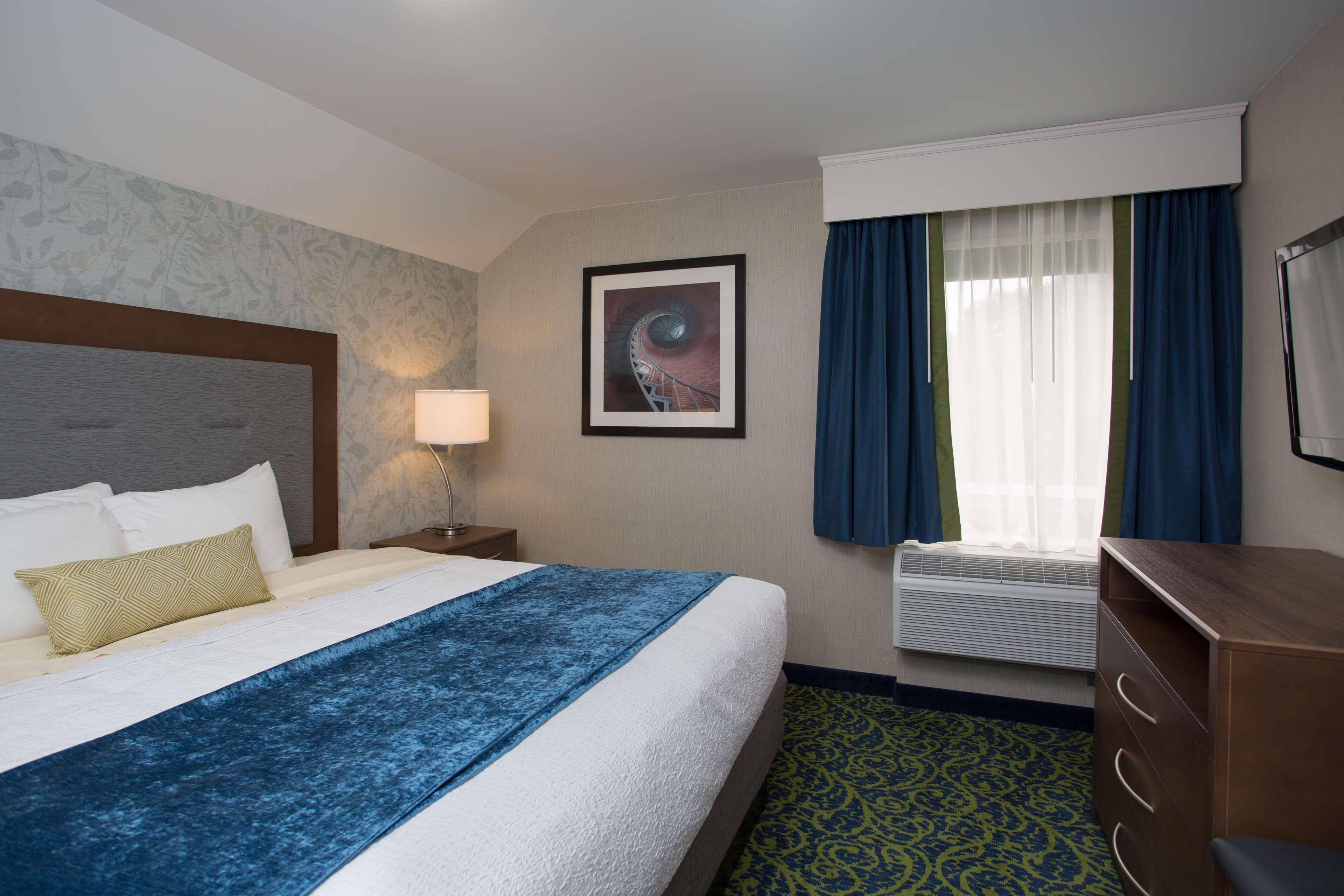 A hotel room with a large bed, a nightstand with a lamp, a framed picture on the wall, a window with curtains, and a dresser.