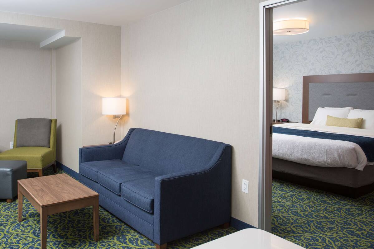 Una habitación de hotel con un sofá azul, una pequeña mesa de madera, un sillón verde y una cama con cabecera.