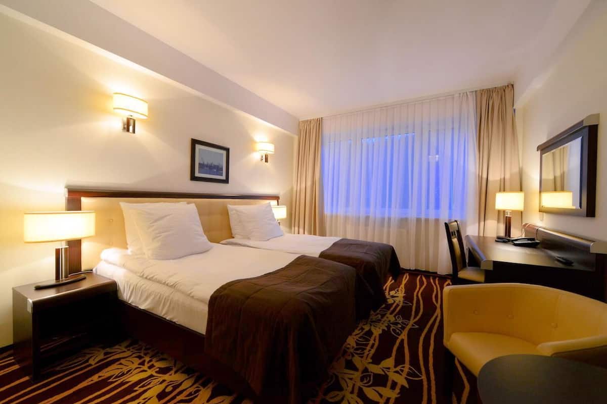 A hotel room with a large bed, two bedside tables with lamps, a desk with a chair, and a large window with curtains.