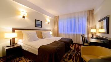 Quarto de hotel com uma cama grande, duas mesas de cabeceira com luminárias, uma escrivaninha com cadeira e uma janela ampla com cortinas.