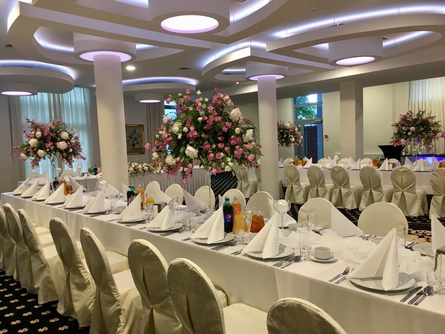 Un salón de banquetes con largas mesas dispuestas para un evento formal, con manteles blancos, sillas ordenadas y grandes centros de flores.