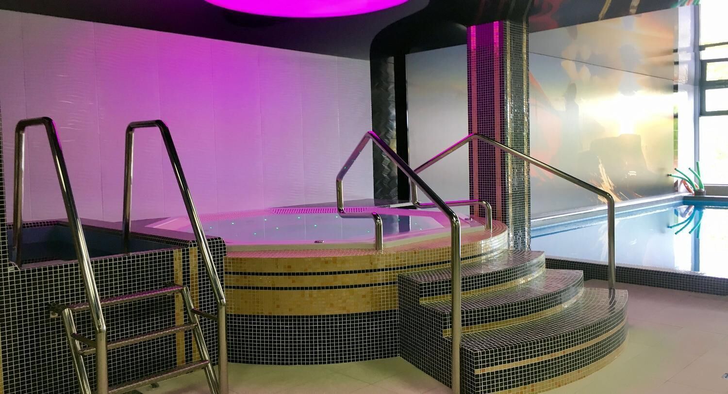 Un espace piscine intérieur moderne, doté d’un grand jacuzzi central, entouré de marches carrelées et de rampes en métal.