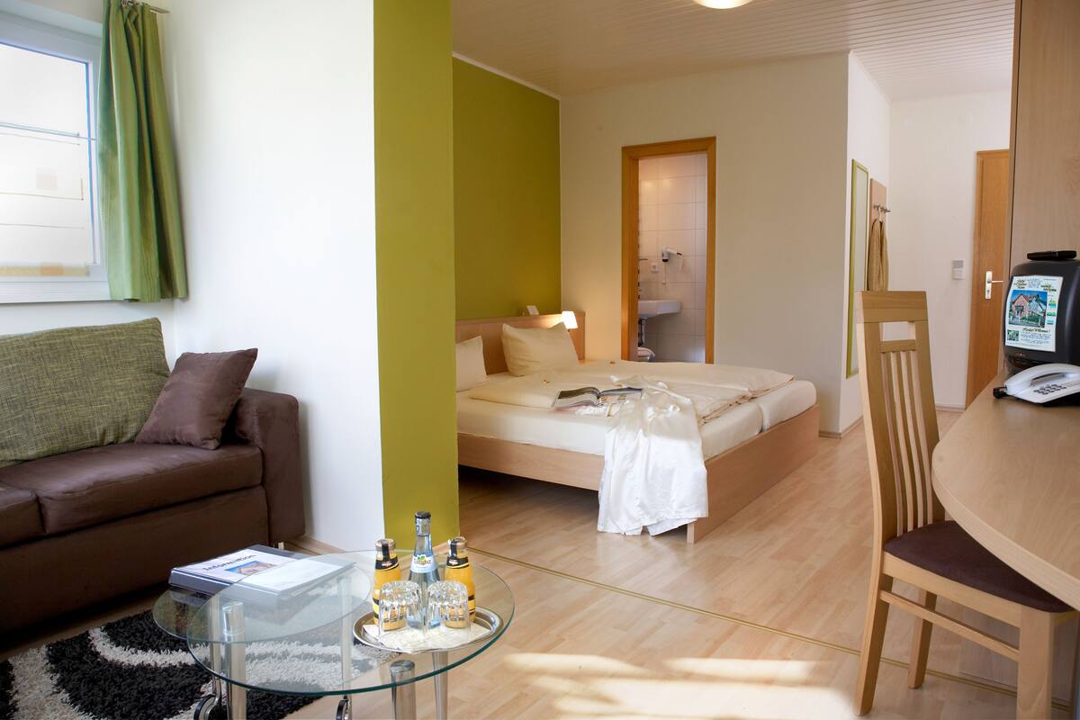 A hotel room with a bed, a sofa, a glass coffee table with bottles, a wooden chair, and a small table with a phone.