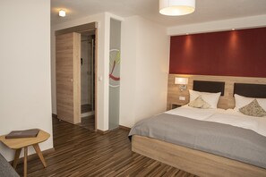 Une chambre d’hôtel moderne avec un plancher en bois, un lit recouvert d’une couverture grise, une table de chevet avec un ordinateur portable, et un mur d’accent rouge.