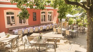 An outdoor dining area with tables and chairs, a building with a red facade, and a tree providing shade.