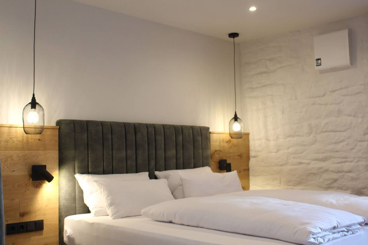 A neatly made bed with white linens, a wooden headboard, and two pendant lights.
