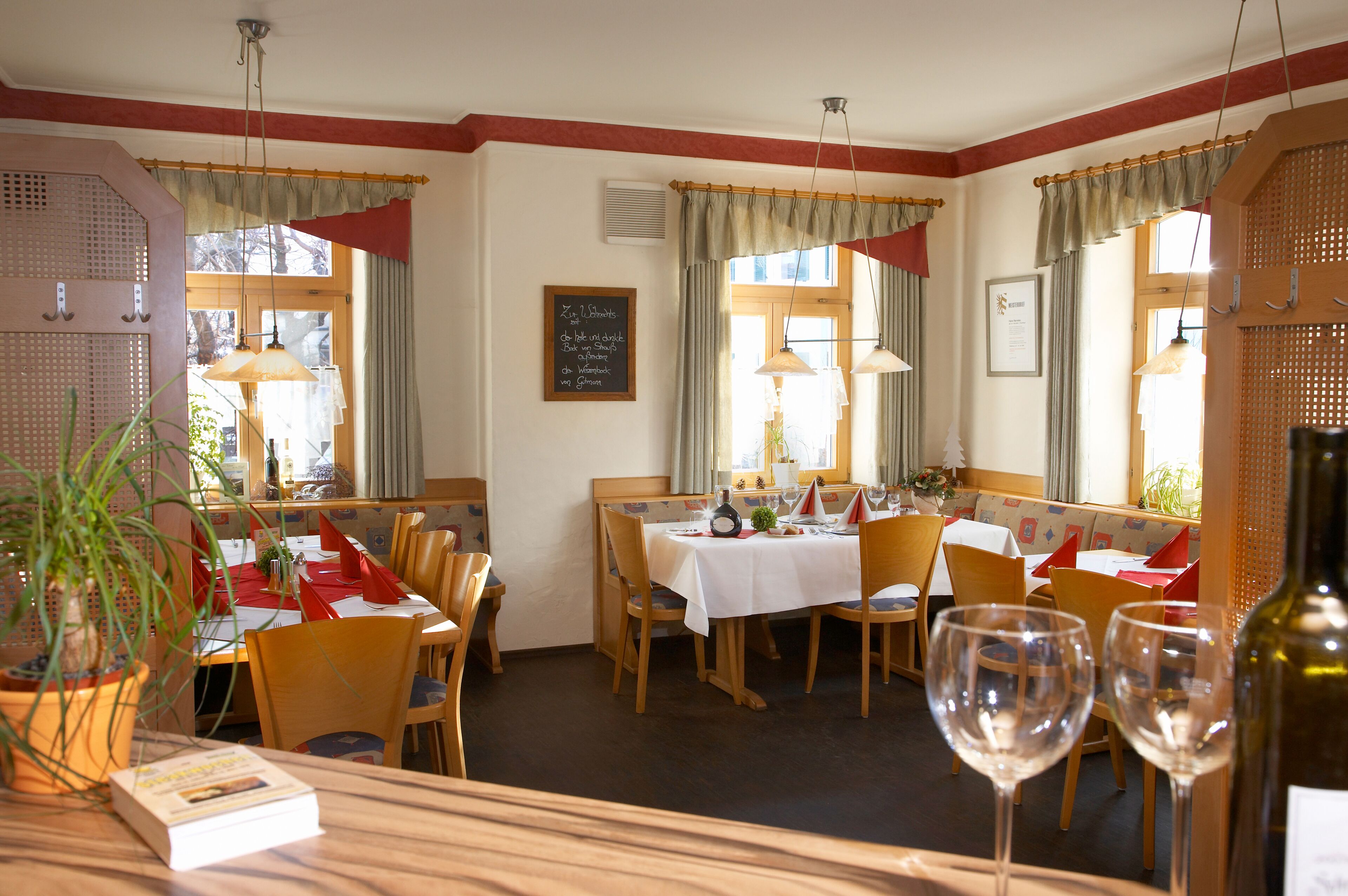 Un restaurante con mobiliario de madera, manteles blancos y servilletas rojas. Hay copas de vino sobre la mesa y una planta en maceta a la izquierda.
