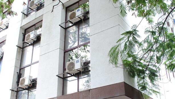 Un edificio de varios pisos con un letrero en idioma extranjero, unidades de aire acondicionado y una persona de pie en la entrada.