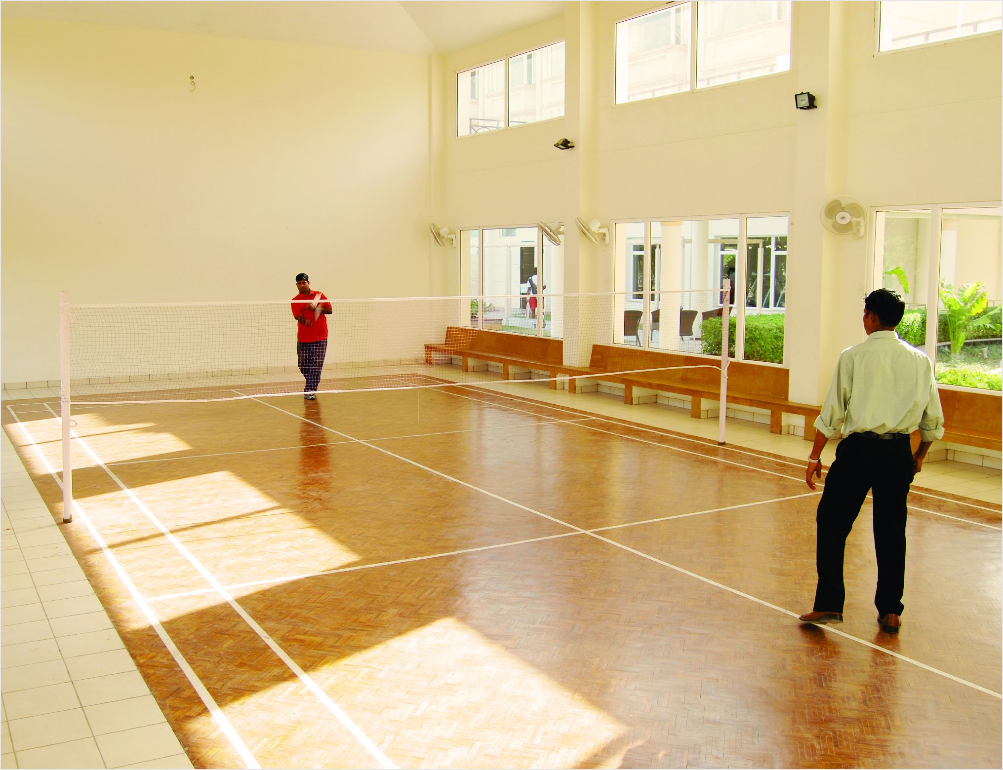 An indoor tennis court with a net, benches, and large windows.