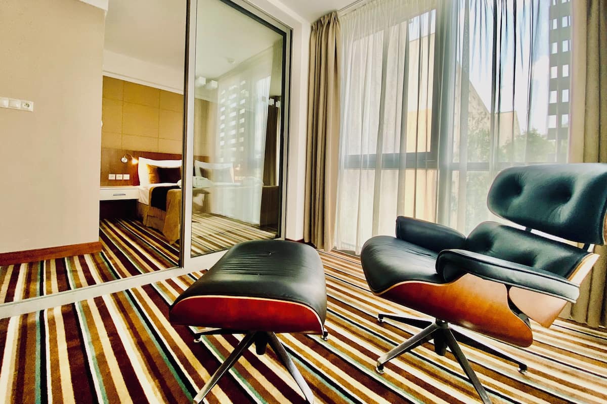 A hotel room with a striped carpet, a black leather chair, a red cushioned bench, and a large window with sheer curtains.