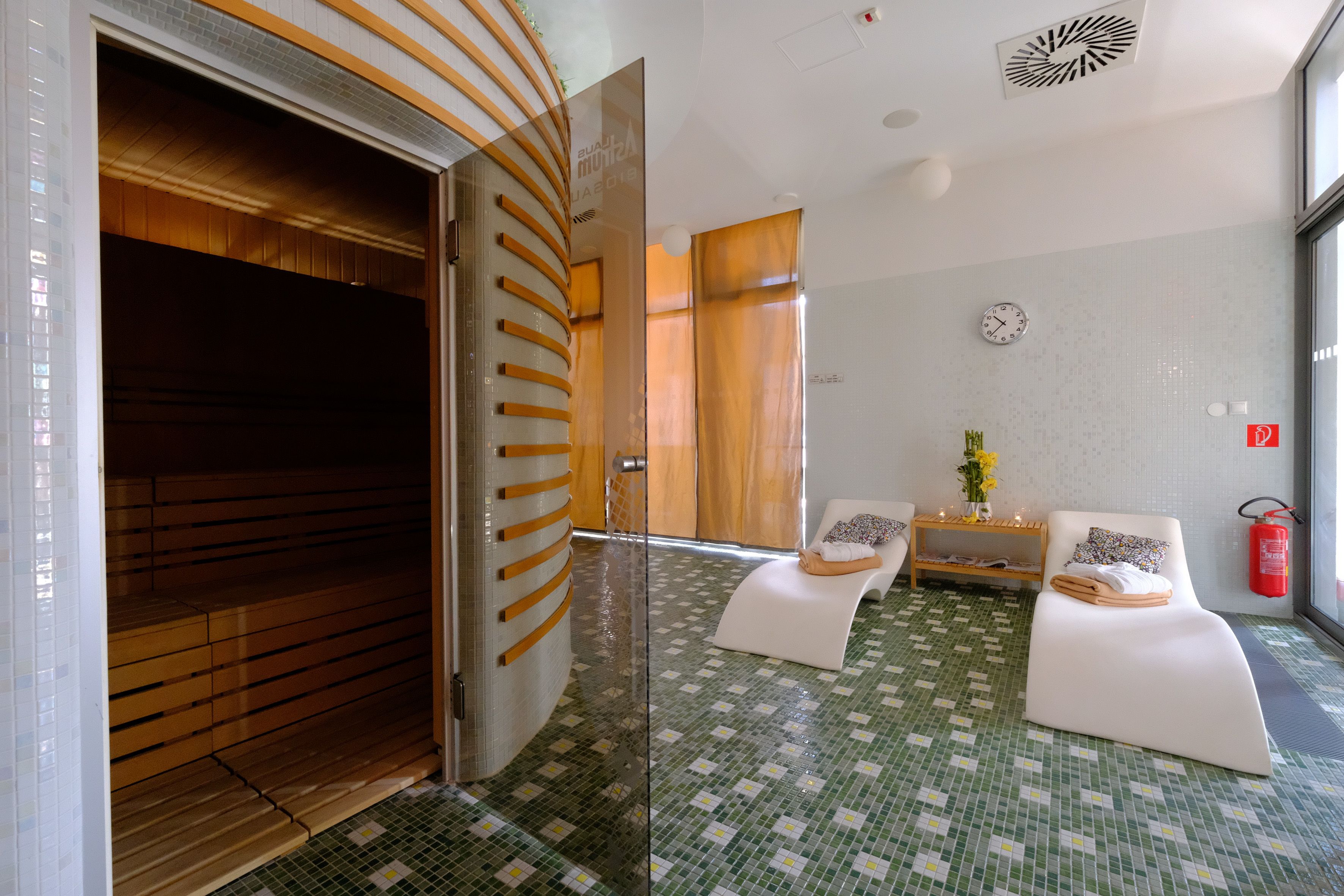 Ett modernt spa-rum med en glasdörr som leder till en bastu, en receptionsdisk och en sittgrupp med en klocka och en brandsläckare på väggen.