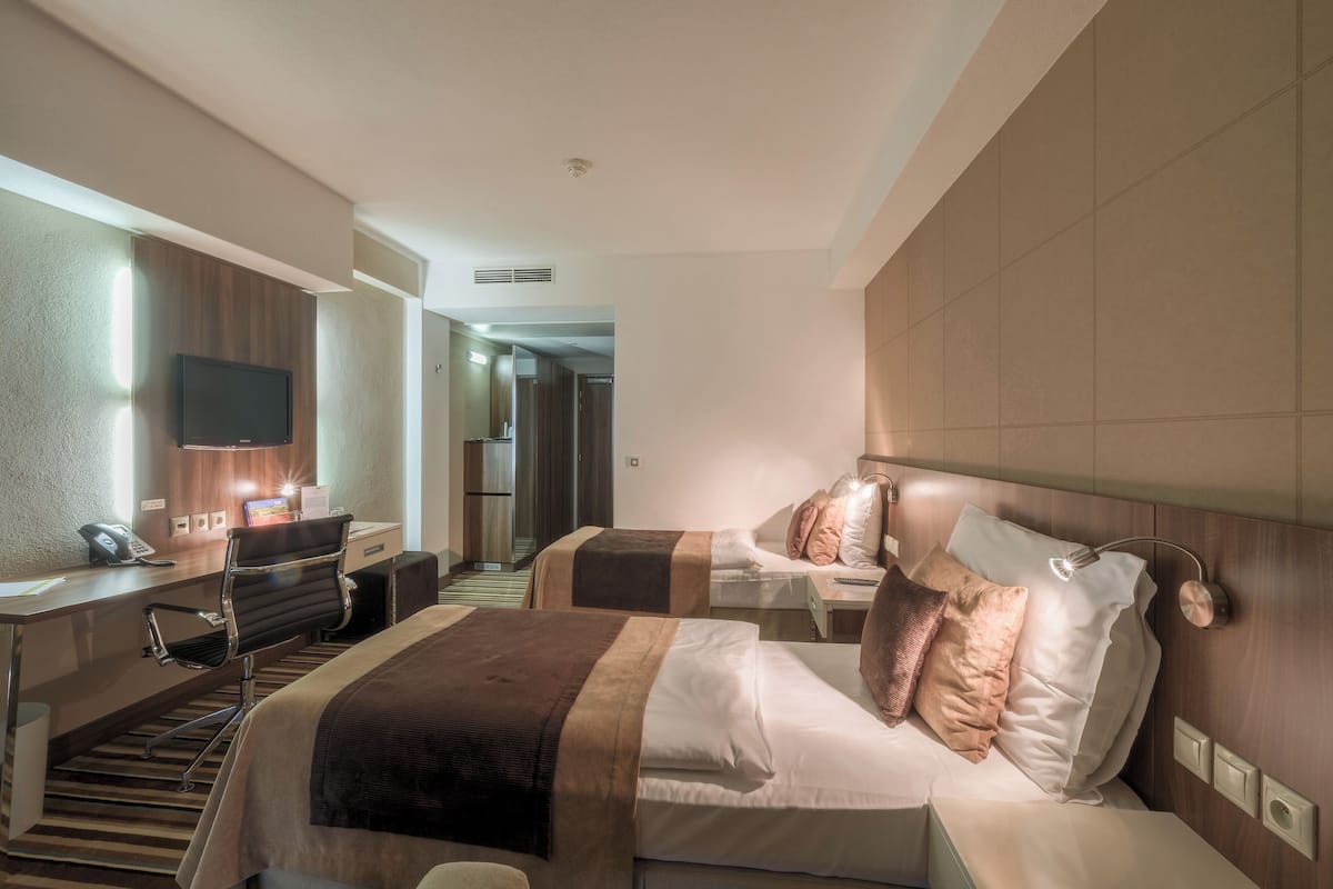 A hotel room with two beds, a desk, a chair, a TV, and a bathroom visible in the background.