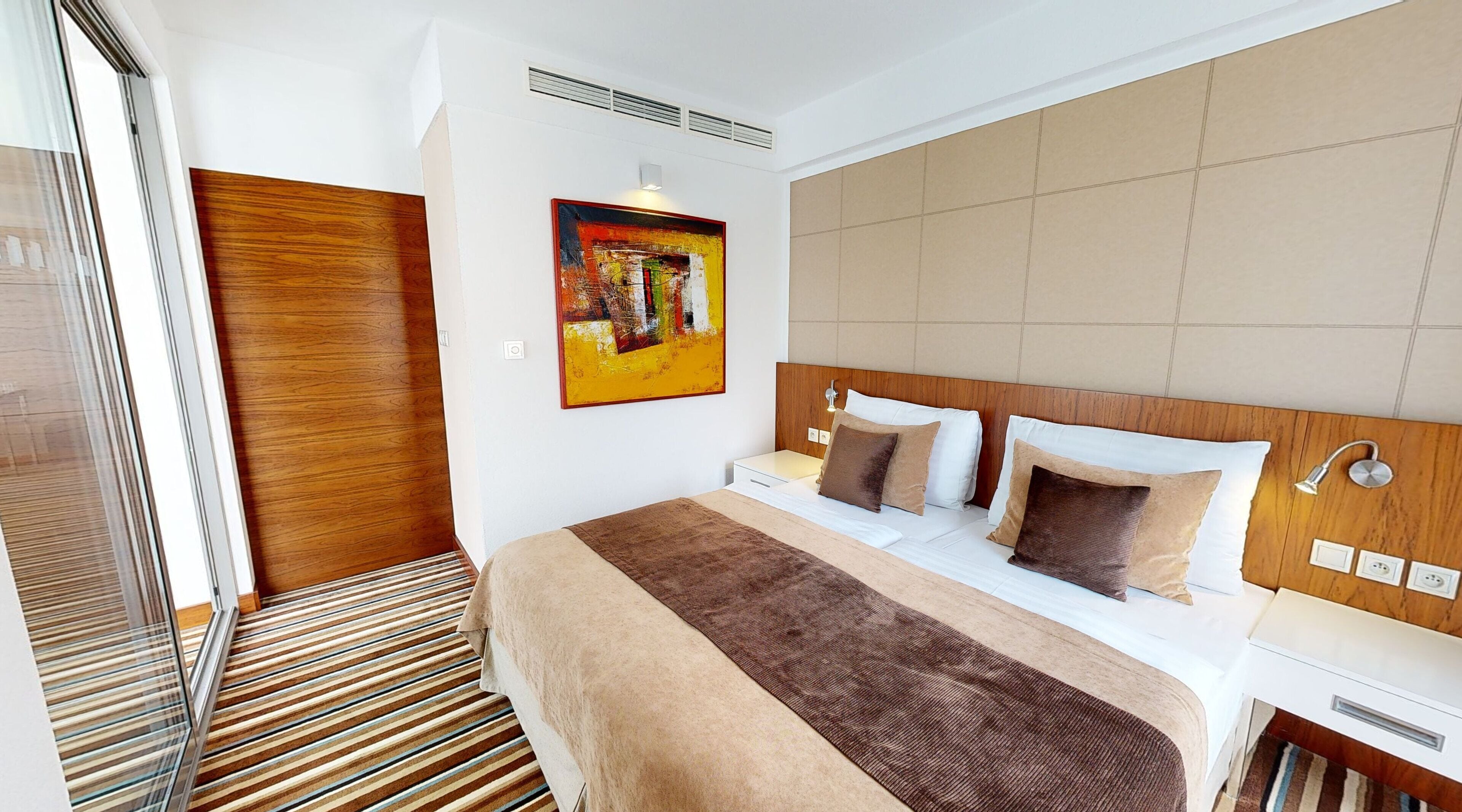 Una habitación de hotel moderna con una cama grande, un cabecero de madera, un cuadro en la pared y una alfombra a rayas.