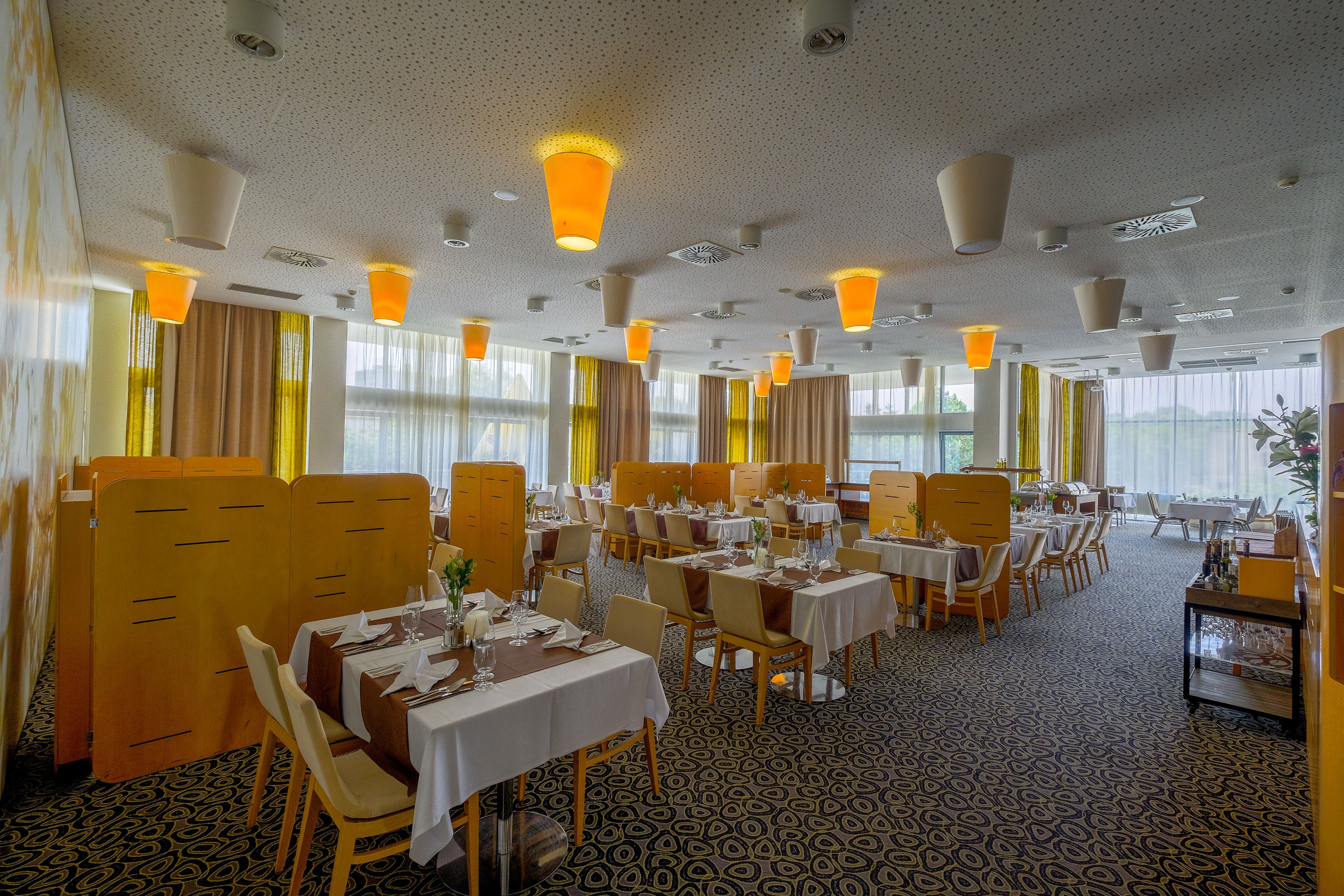 Een restaurantgedeelte in een hotel, met gedekte tafels, grote ramen met gordijnen en plafondlampen.