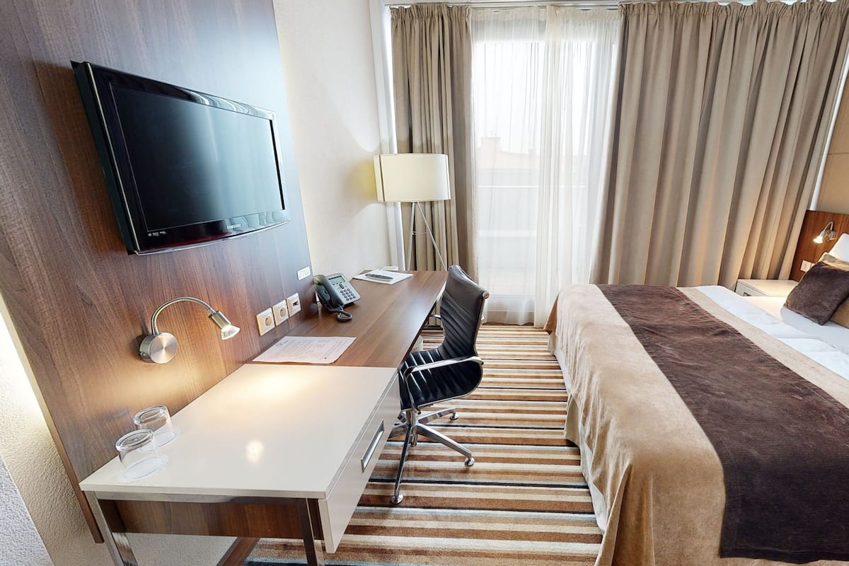 A hotel room with a bed, a desk with a chair, a TV mounted on the wall, a lamp, and a window with curtains.