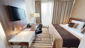 A hotel room with a bed, a desk with a chair, a TV mounted on the wall, a lamp, and a window with curtains.