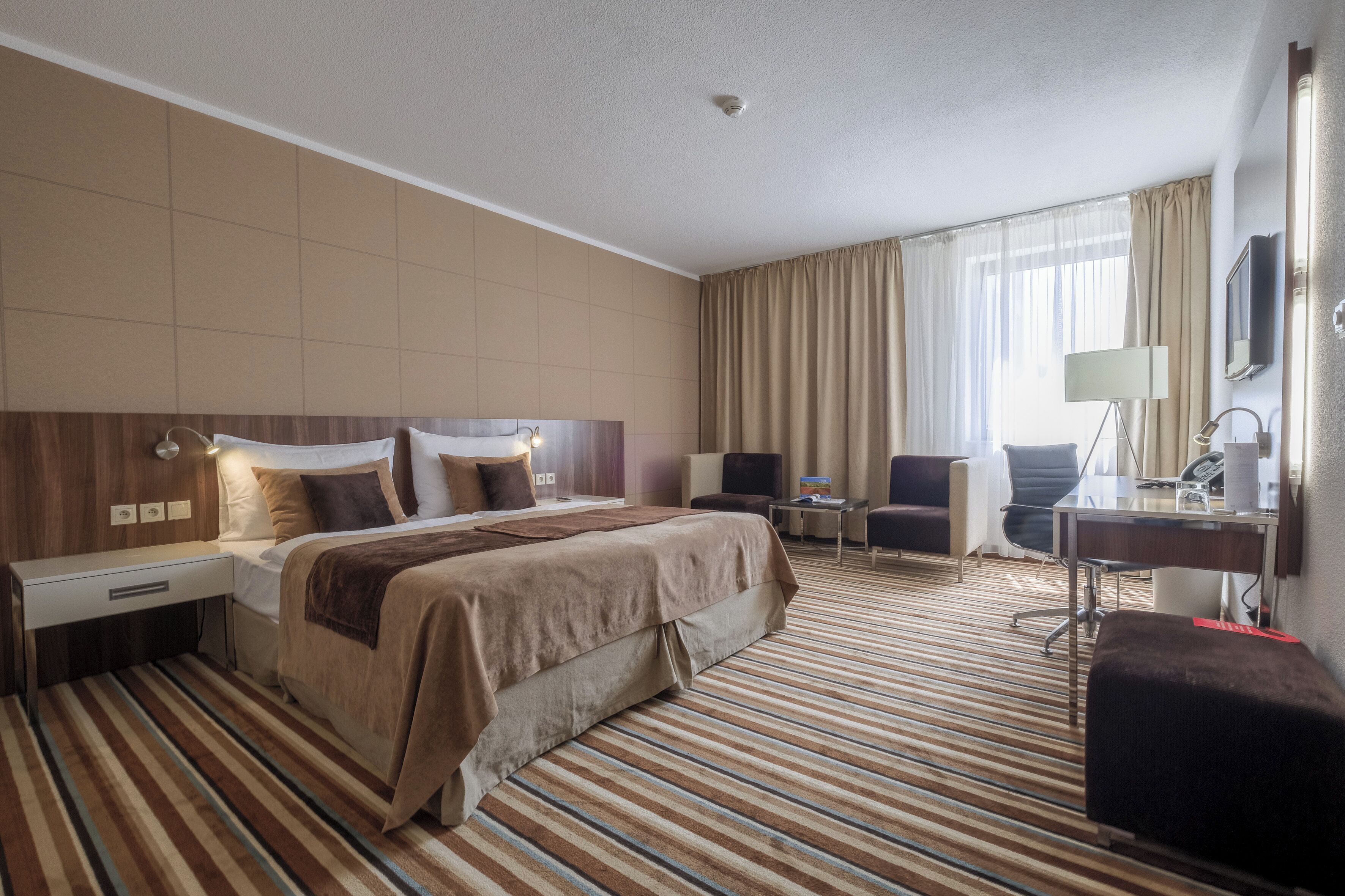 A hotel room with a large bed, a desk, a chair, a TV, and a window with curtains.