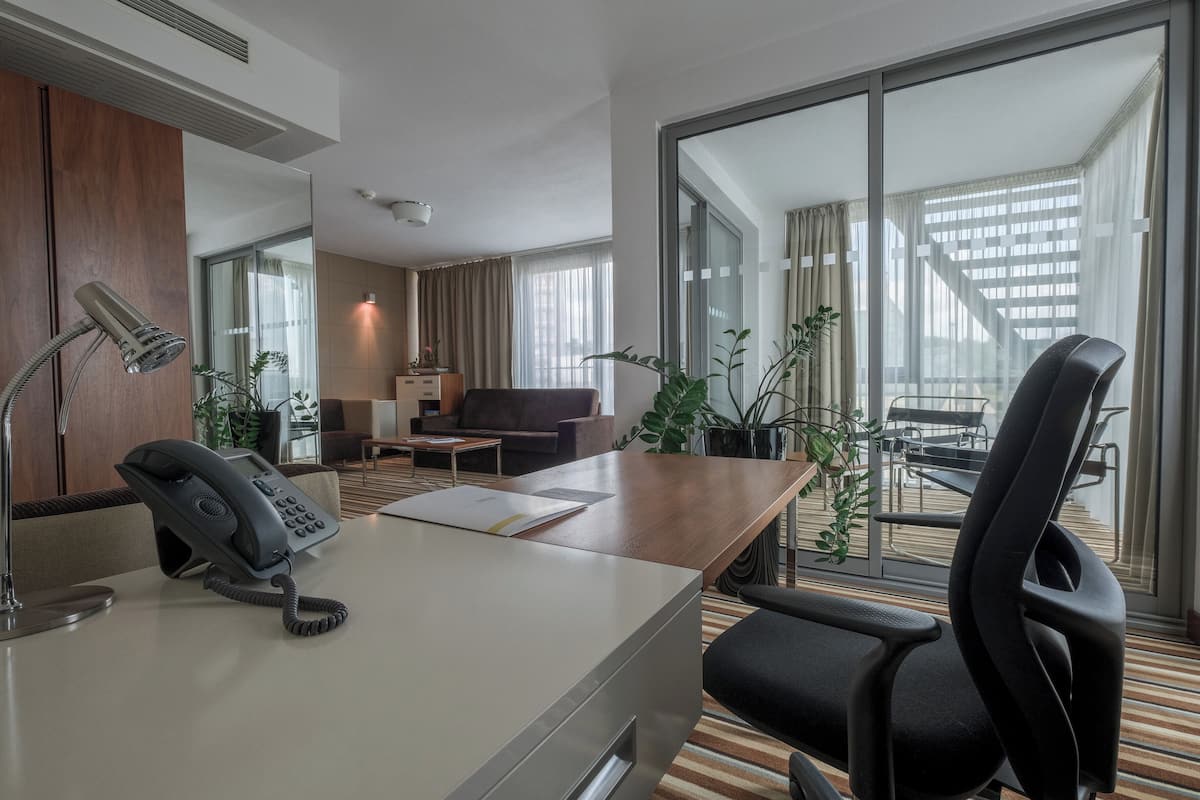 A hotel room with a desk, phone, and chair, a sofa, a coffee table, and large windows with a view of a building.