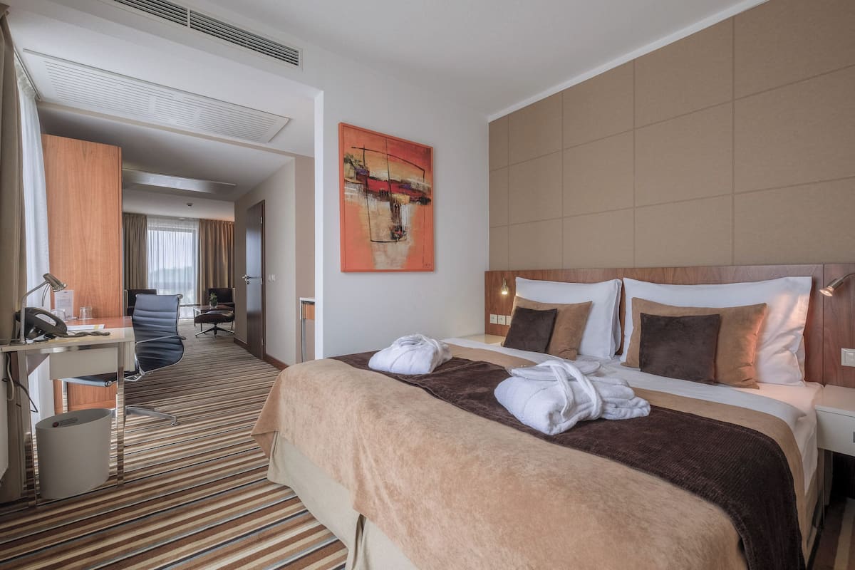 A hotel room with a large bed, a desk, and a chair. There is a painting on the wall and a window with curtains.