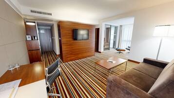 A hotel room with a striped carpet, a wooden wardrobe, a TV mounted on the wall, a sofa, a small table, and a desk with a chair.