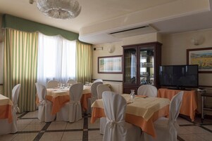 A dining area with tables set for six, each with white chairs and orange tablecloths. There is a television on a stand to the right and a cabinet with glass doors to the left.