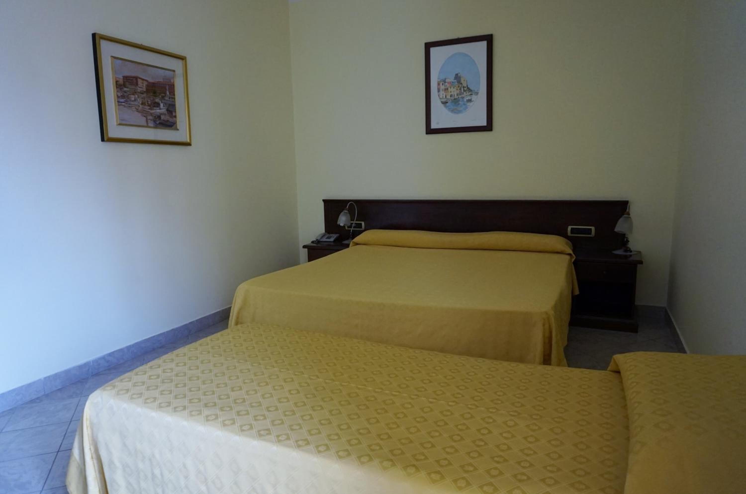 A hotel room with two beds, a wooden headboard, a nightstand with a lamp, and two framed pictures on the wall.