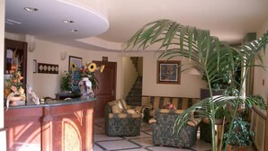 A lobby with a reception desk, two patterned sofas, a large potted plant, and a staircase in the background. - Hotel Ristorante Stella 2000 (Olbia)