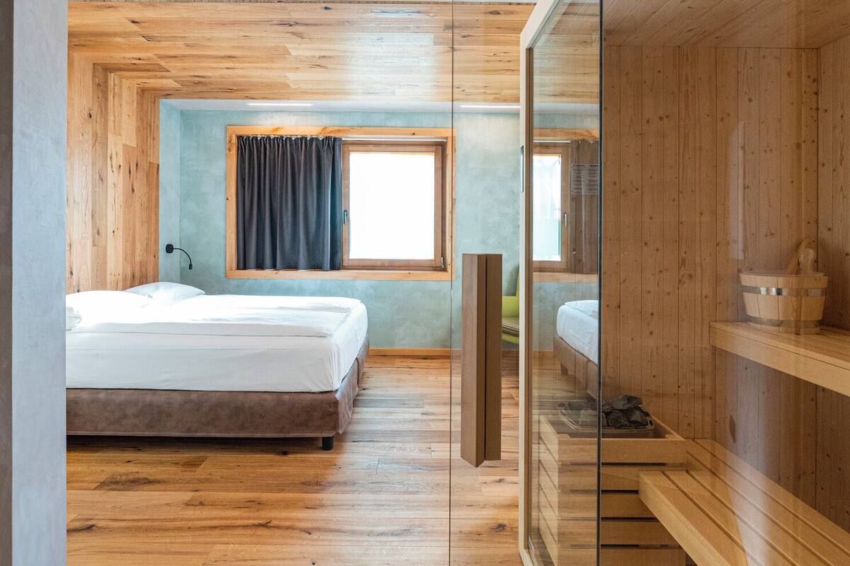 A modern bedroom with two beds, wooden flooring, a large window with curtains, and a sauna area.