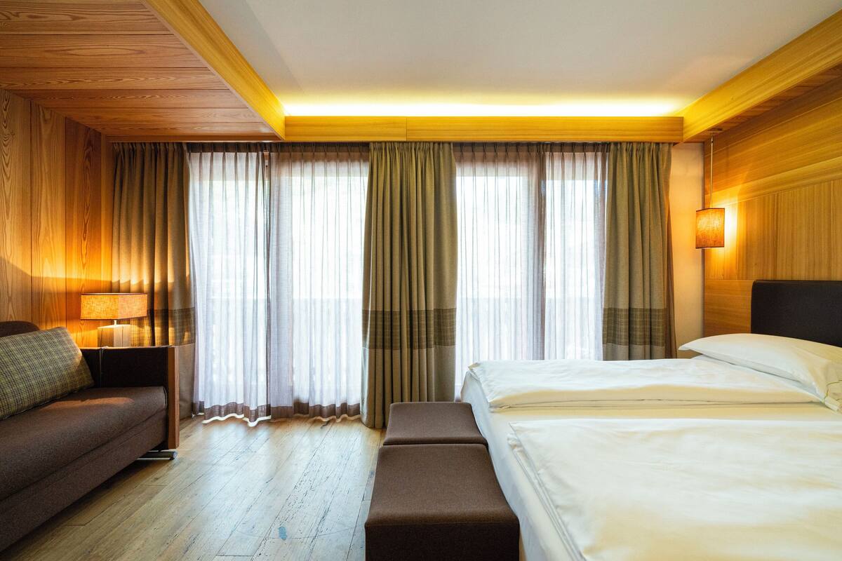 A hotel room with a wooden ceiling, a bed with white linens, a brown sofa, a bench, and curtains.