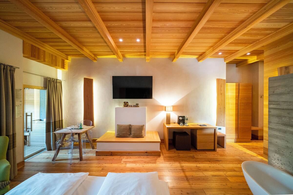 A spacious room with wooden flooring, a flat-screen TV mounted on the wall, a seating area with a table and chairs, and a bed with white linens.