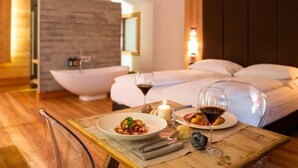 A dining table with plates of food and wine glasses, a candle, and a chair in a hotel room with a bed and a bathtub in the background.
