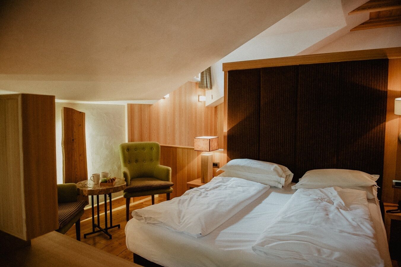A hotel room with a large bed, a green armchair, a small table with a lamp, and wooden paneling on the walls.