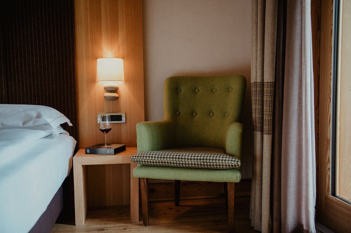 A hotel room with a neatly made bed, a green armchair with a patterned cushion, a bedside table with a wine glass and a digital clock, and a wall-mounted lamp.