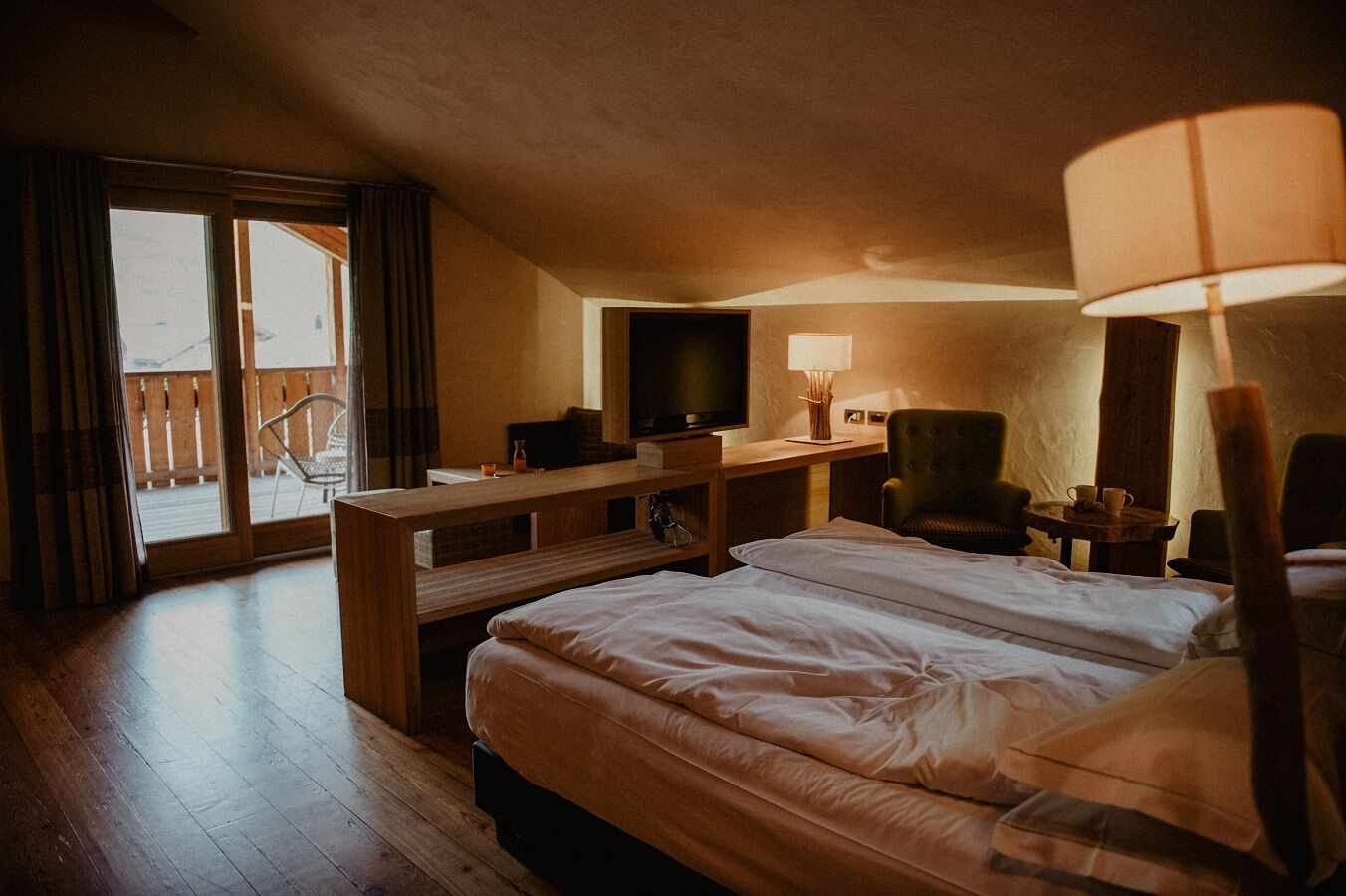 A hotel room with a bed, desk, chair, TV, lamp, and a balcony with curtains.