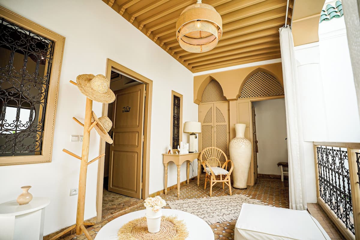 A room with a wooden ceiling, a woven ceiling light, a straw hat rack, a wicker chair, a white vase with flowers, and a patterned rug.