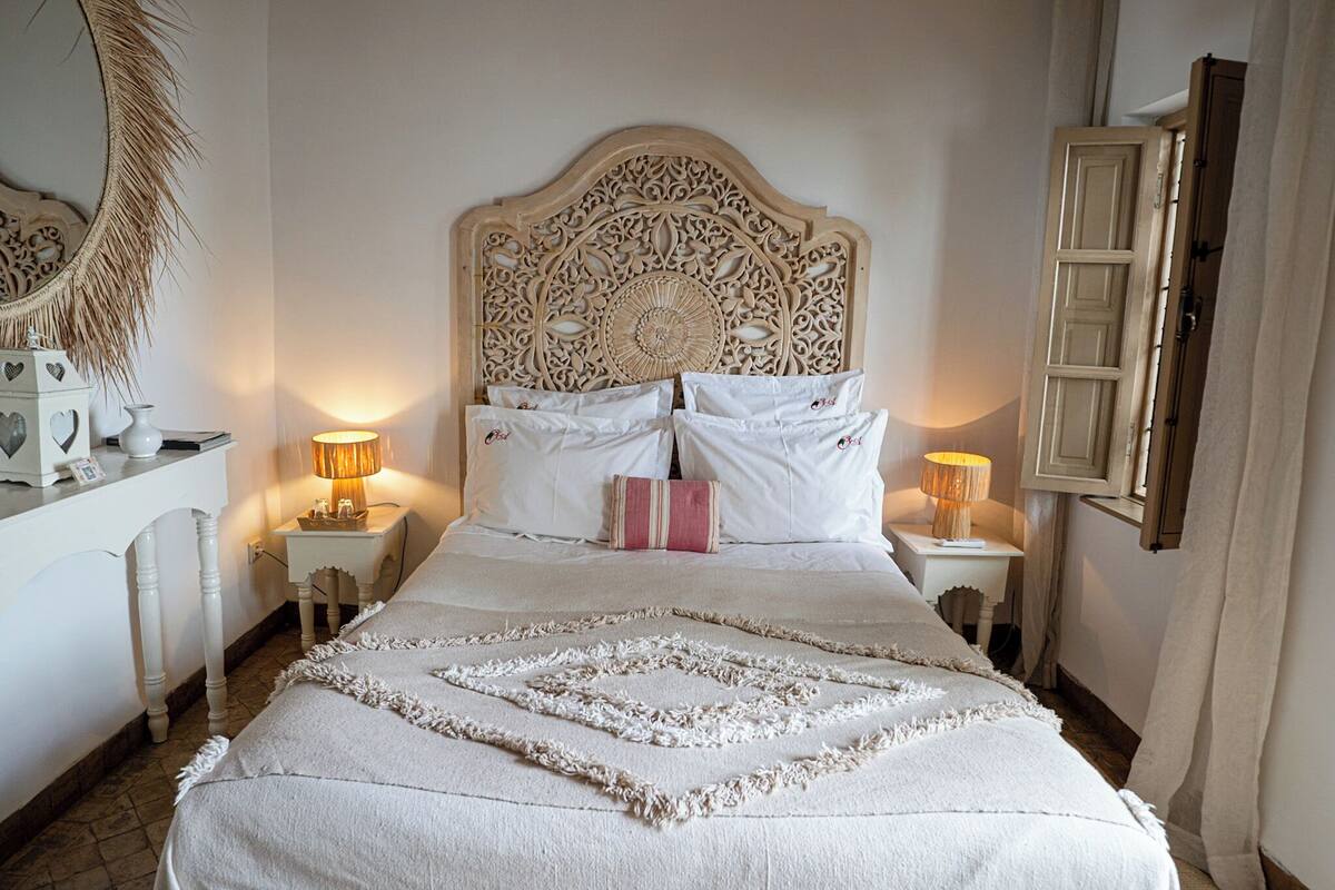 A neatly arranged bedroom with a large bed, two bedside tables with lamps, a decorative headboard, and a mirror on the wall.