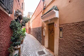 Un estrecho callejón empedrado flanqueado por edificios, con una puerta que tiene un letrero, un ventanal pequeño y un toldo decorativo.