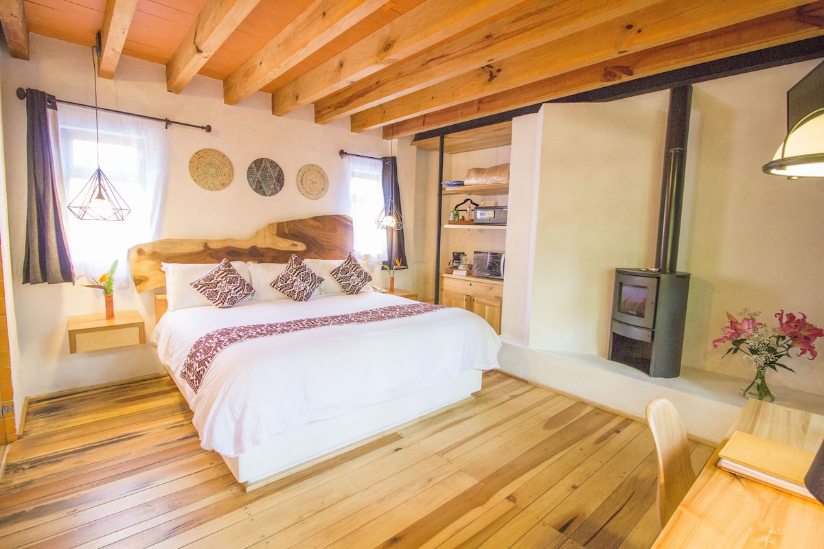 A bedroom with a wooden bed, wooden floors, a wooden ceiling, a window with curtains, a bedside table with a plant, and a wall with decorative plates.